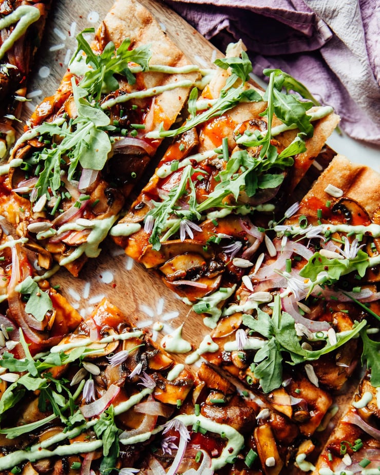 An overhead shot of a grilled flatbread with mushrooms, red onions, BBQ sauce, and a drizzle of a pale green chive sauce. The flatbread is cut into square pieces and is garnished with fresh arugula.