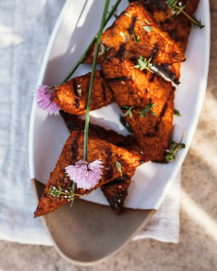 An overhead shot of marinated and grilled tempeh in direct lighting. The pieces of tempeh are garnished with chive flowers.