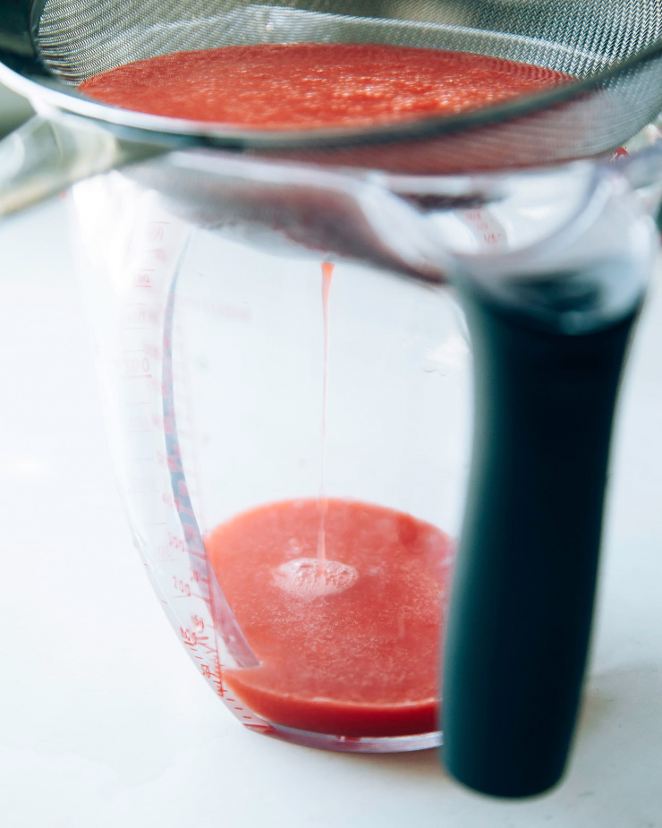 Displays stawberry, watermelon, and lime juice being run through a fine mesh strainer.