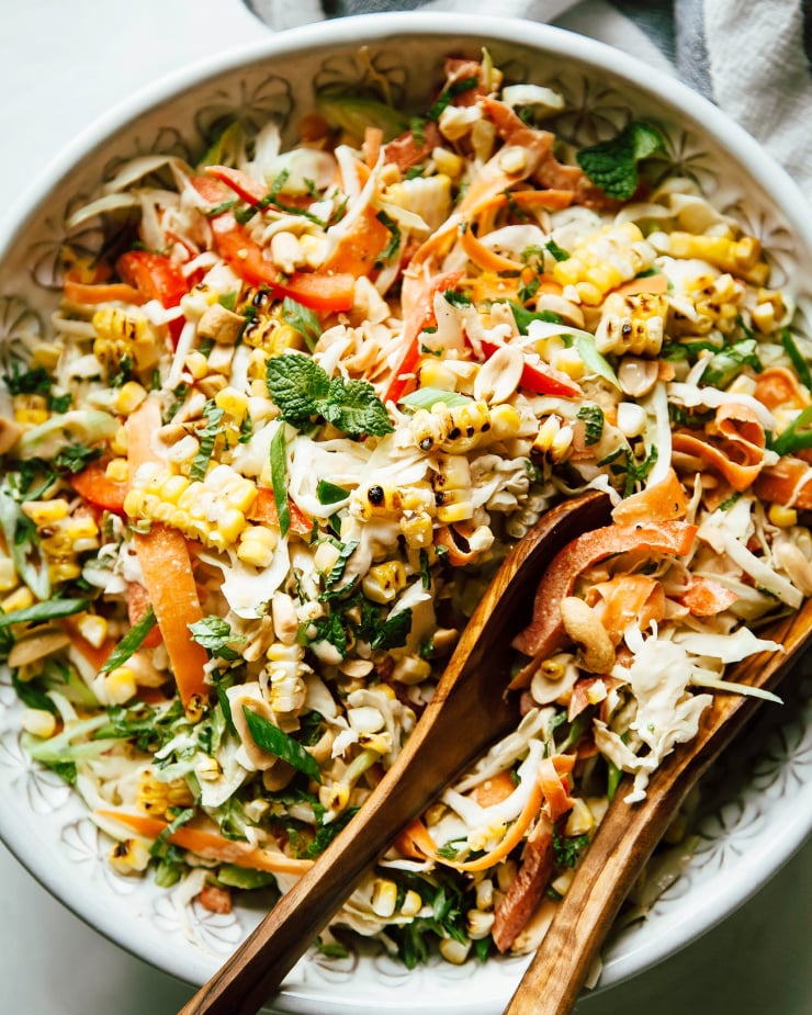 An up close, overhead shot of charred corn slaw with peanut butter lime dressing. The slaw is served in a white patterned bowl with wood serving tongs.