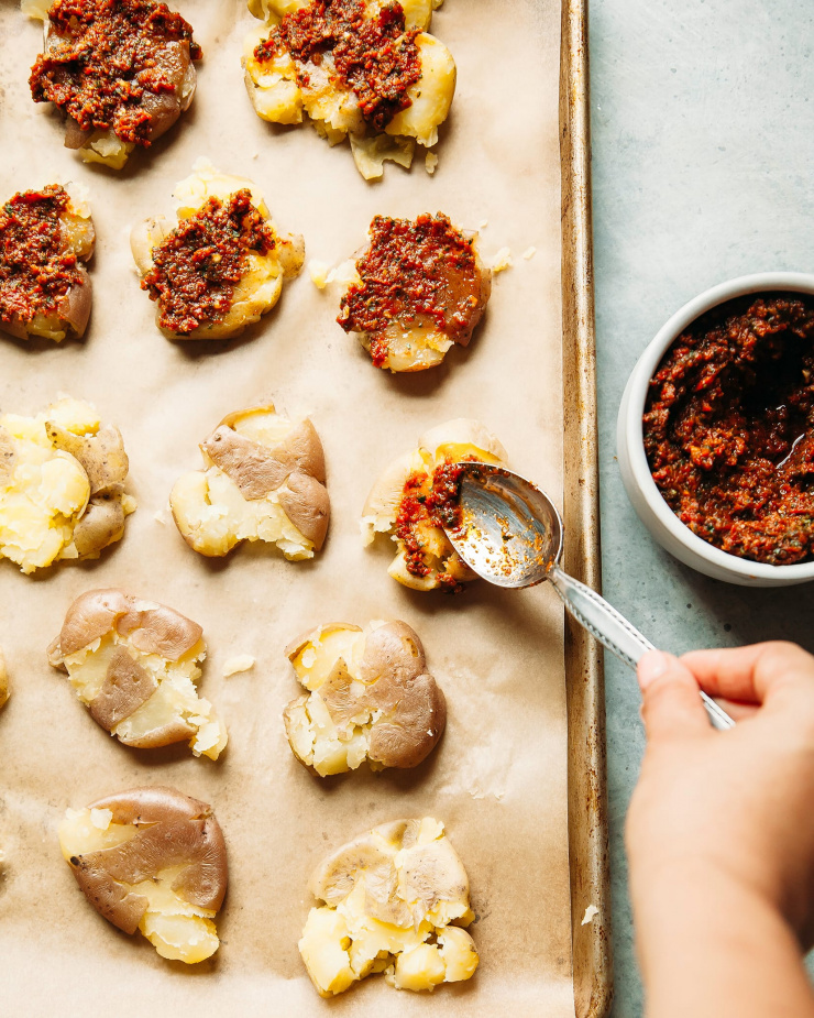 A hand is spooning pesto on top of cooked and smashed down mini potatoes.