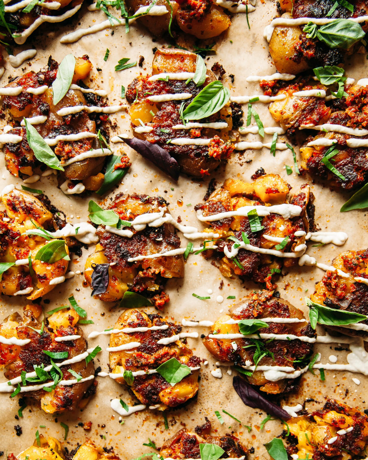 Overhead shot of smashed sun dried tomato pesto potatoes with a creamy vegan Caesar drizzle over the top. The potatoes are also garnished with fresh basil leaves.