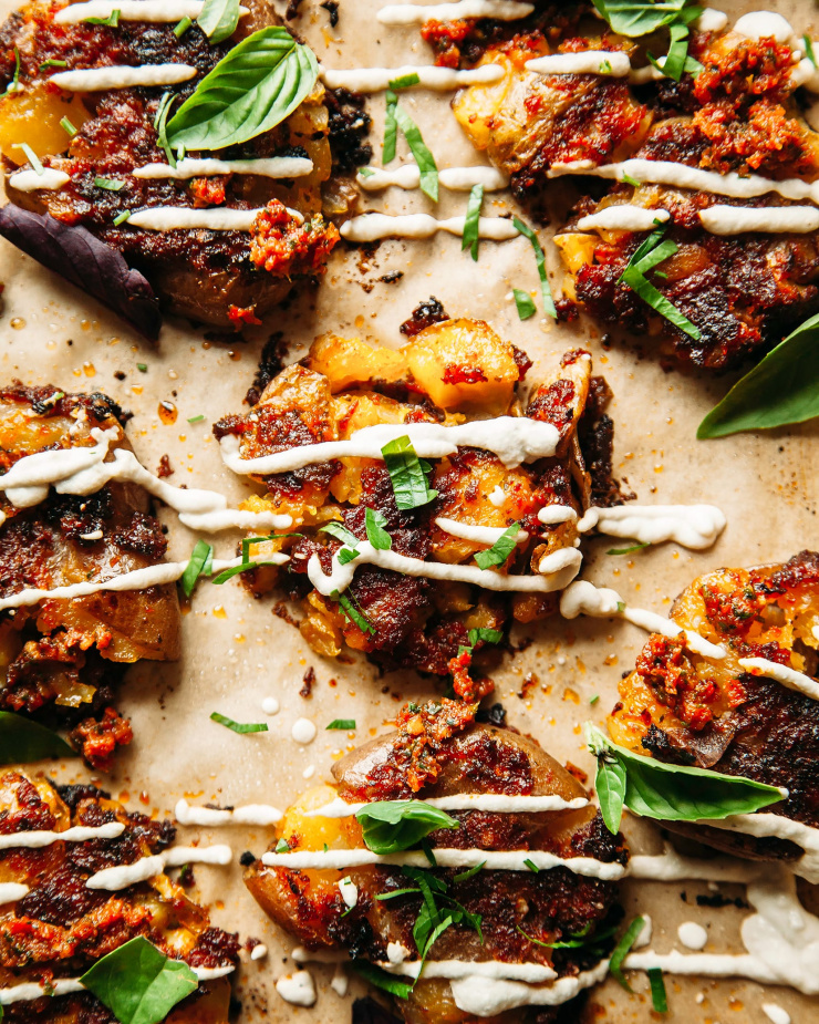 Up close, overhead shot of smashed sun dried tomato pesto potatoes with a creamy vegan Caesar drizzle over the top. The potatoes are also garnished with fresh basil leaves.