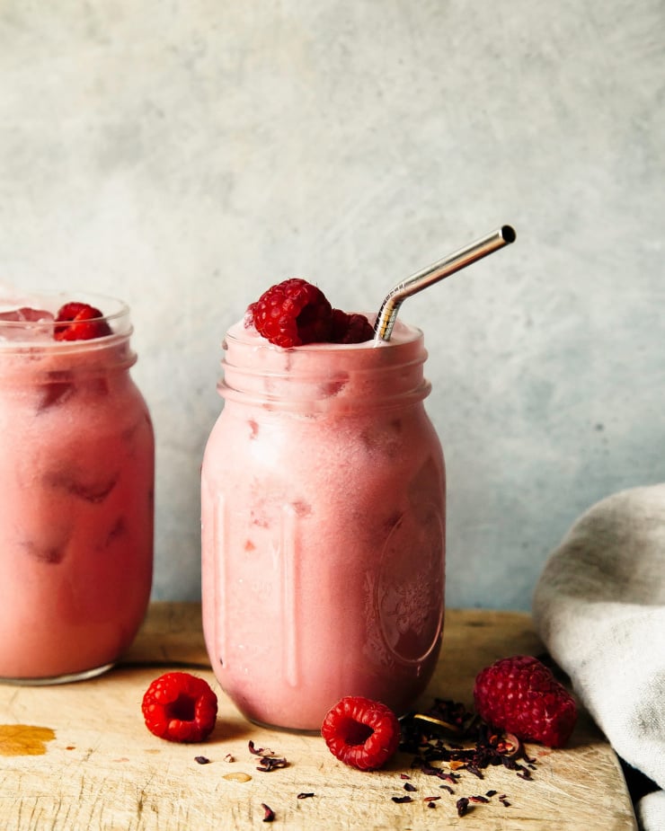 A straight on shot of a light pink raspberry hibiscus pink drink in two small mason jars against a light blue background.
