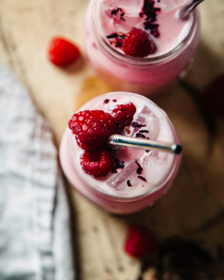 Overhead shot of creamy raspberry hibiscus pink drink in two mason jars on top of a light wood cutting board. The drink is gar in shed with fresh raspberries.