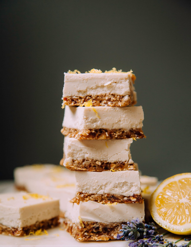 A head-on shot of a stack of creamy vegan lemon bars against a dark grey background. There is a halved lemon and sprigs of lavender nearby.