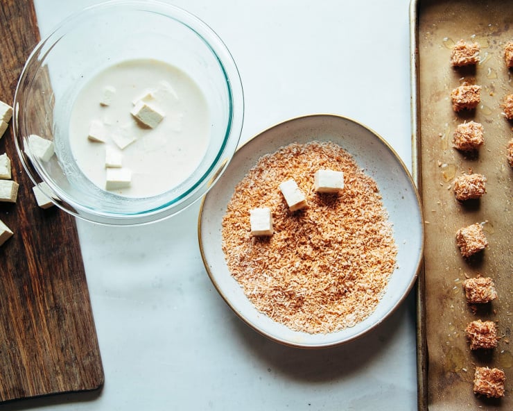 An overhead shot of tofu being coated in toasted coconut and eventually transferred to a baking sheet, all on a white background.