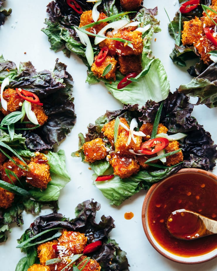An overhead shot of vegan red leaf lettuce wraps containing crispy and golden cubes of coconut coated tofu. A small bowl of a deep red, chili flecked sauce is nearby.