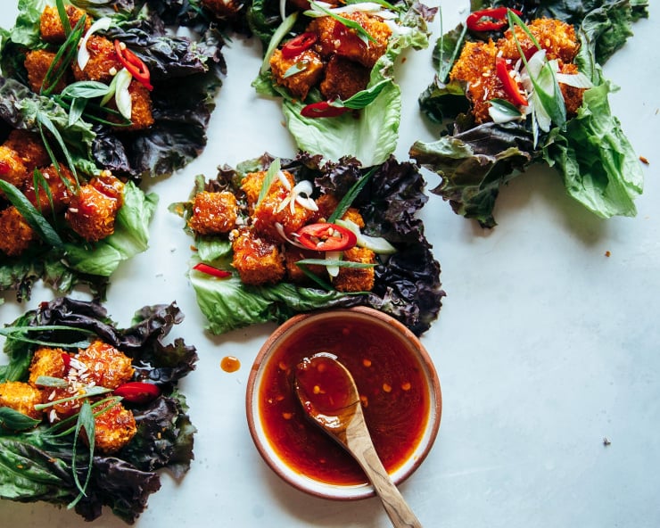 An overhead shot of crispy coconut tofu lettuce wraps with a bowl of sweet chili sauce for drizzling nearby. The tofu is wrapped in red leaf lettuce and the cubes of tofu are deep golden brown.