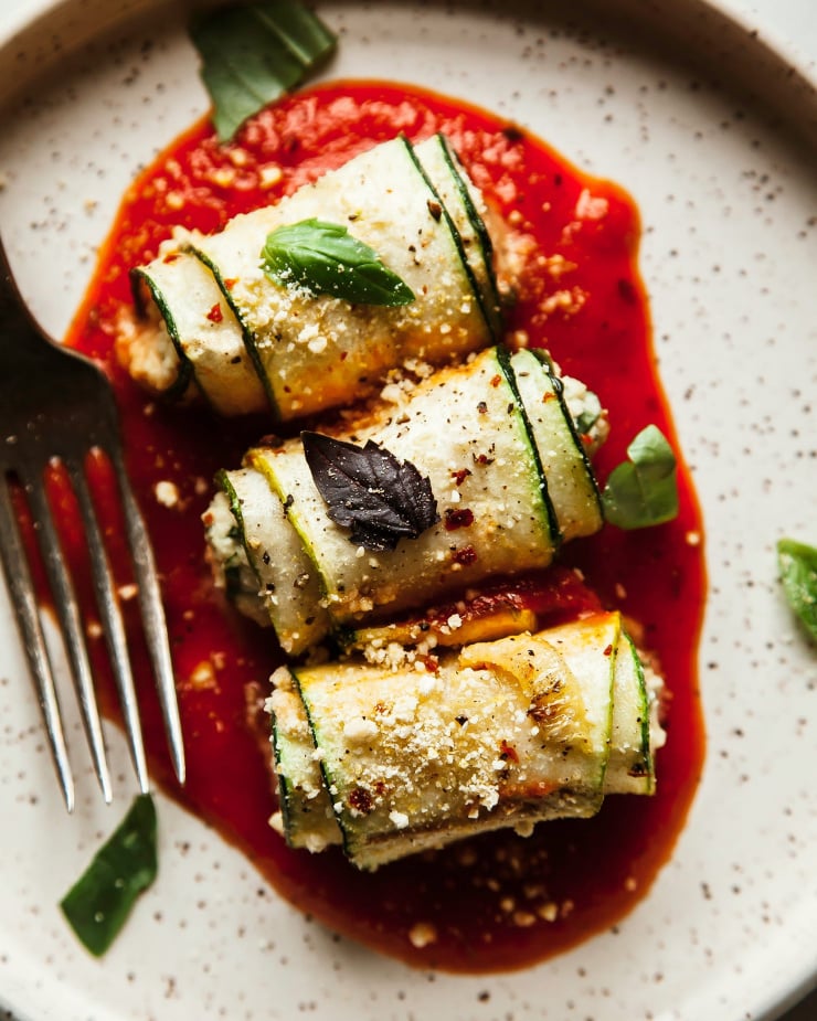 An up close, overhead shot of 3 rolls of zucchini Involtini on a little moat of marinara sauce, plated on a white speckled plate. A fork is to the side.