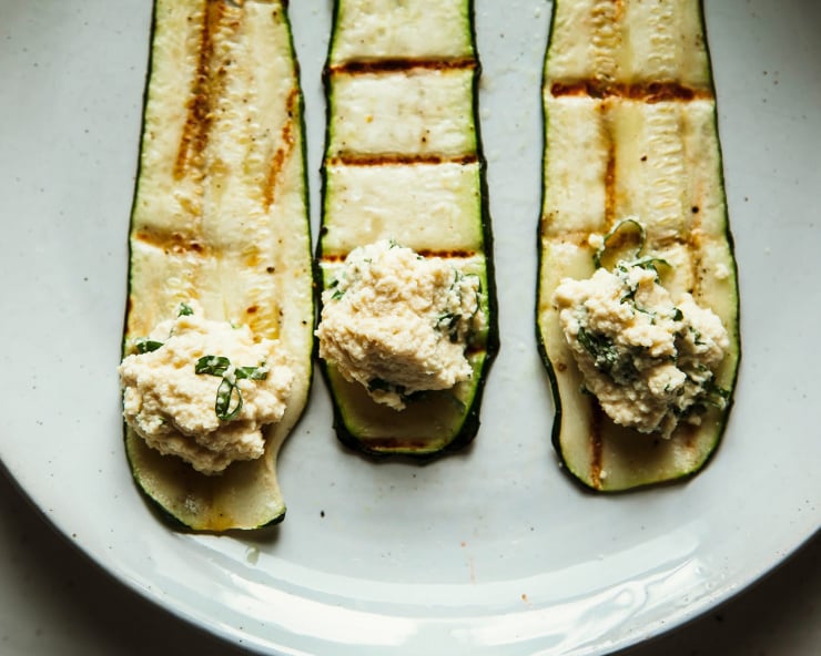 An overhead shot of three grilled strips of zucchini with dollops of herbed almond ricotta on top, ready to be rolled up.