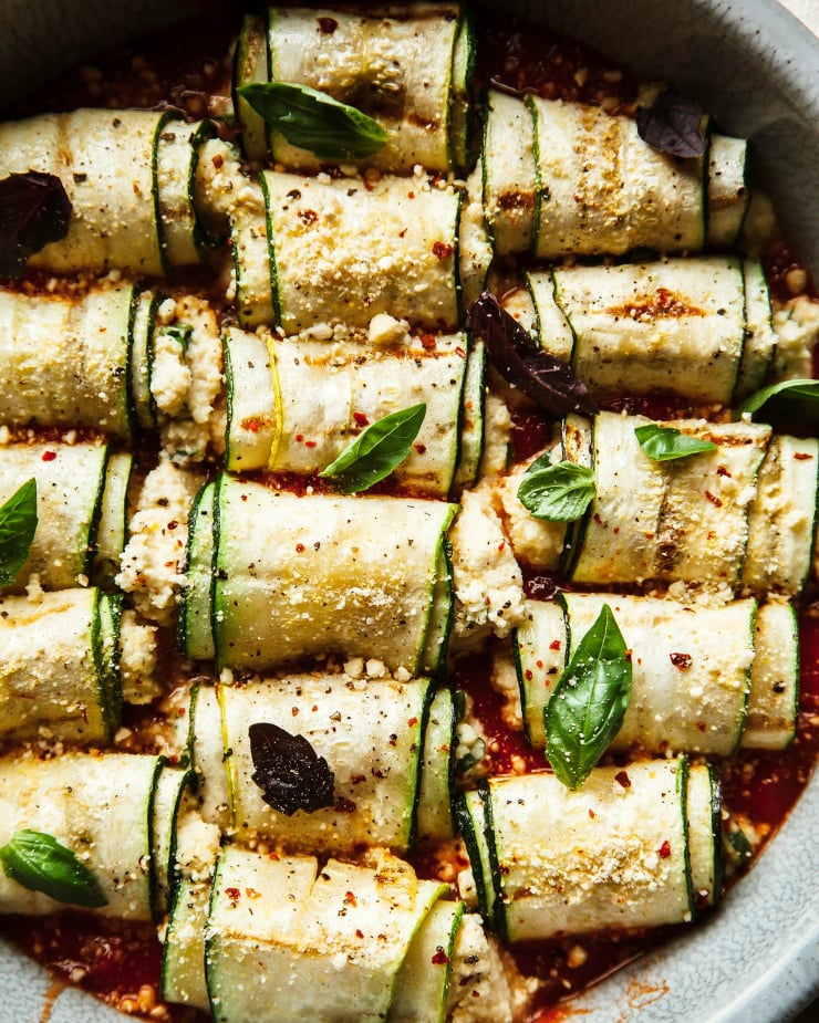 An up close, overhead shot of zucchini Involtini with almond ricotta in a baking dish.The rolls of zucchini are topped with basil leaves.