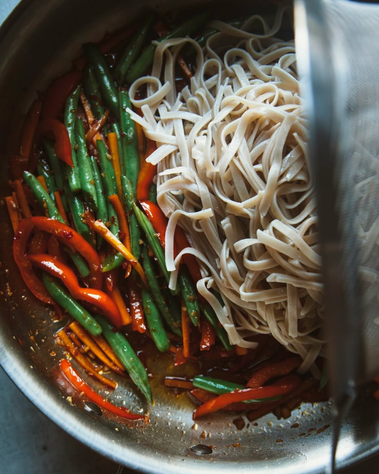 Image shows cooked noodles being added to a pan with sautéed vegetables.