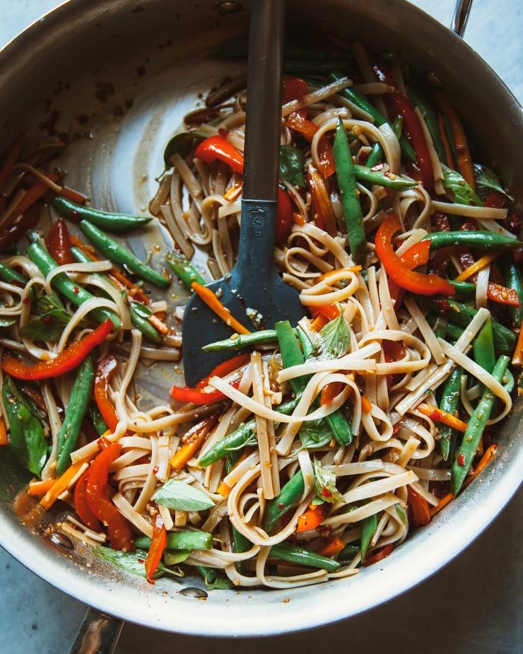 Image shows noodles and vegetables being stirred together in a sauté pan.