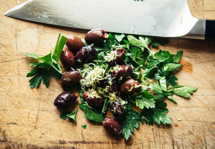 Olives, parsley and lemon zest on a worn wood cutting board, about to be chopped.
