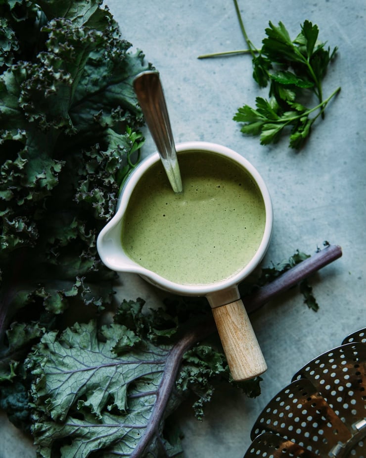 An overhead shot of green pepita lime sauce in a small white pitcher on a blue background. Kale and parsley leaves are nearby.