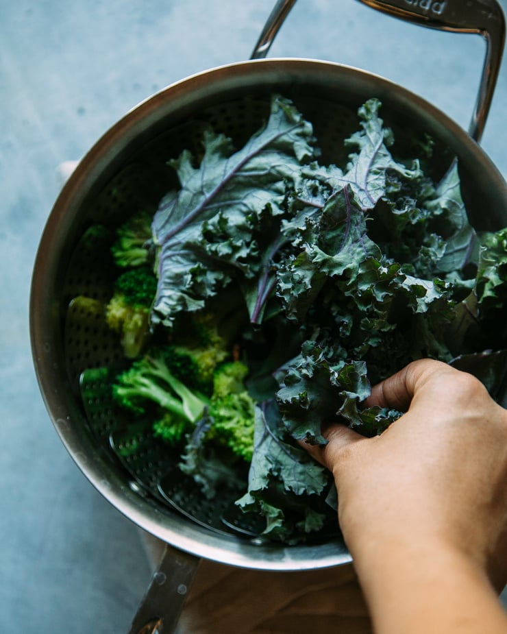 A hand is shown stuffing kale leaves into a stainless steel saucepan for steaming.