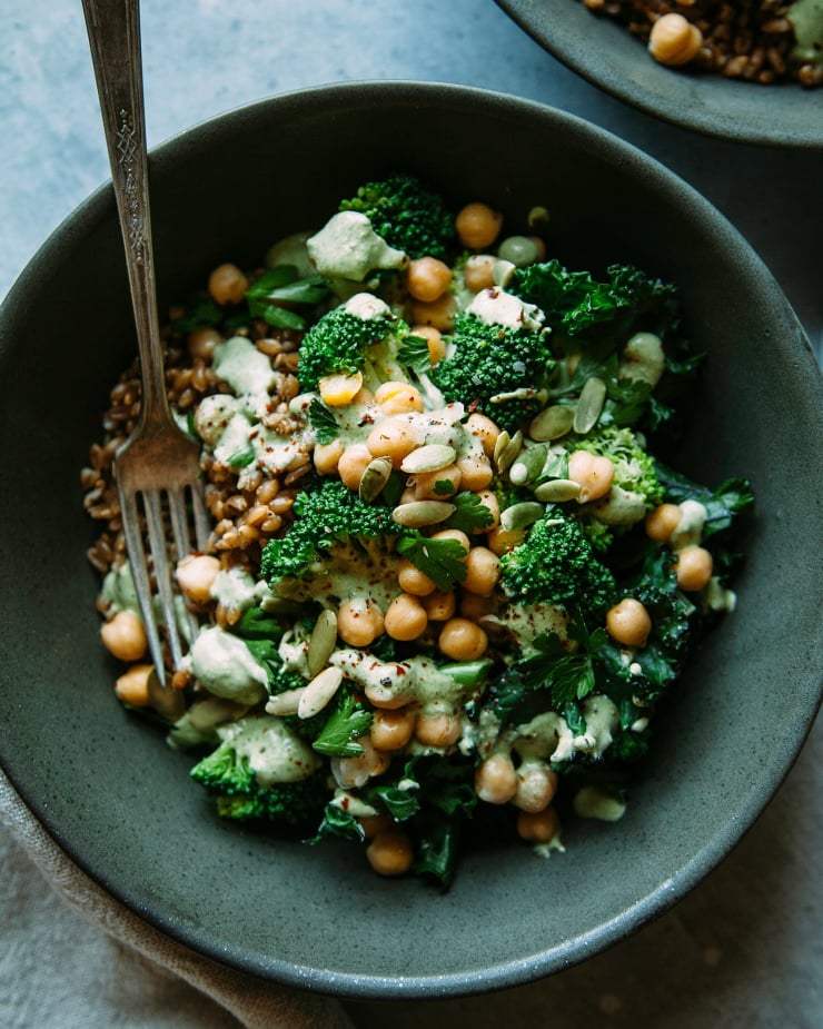 An up close shot of a green goddess bowl with broccoli, kale, chickpeas, farro, and a sage-green pepita lime sauce. The bowl is shown in a deep green matte bowl.