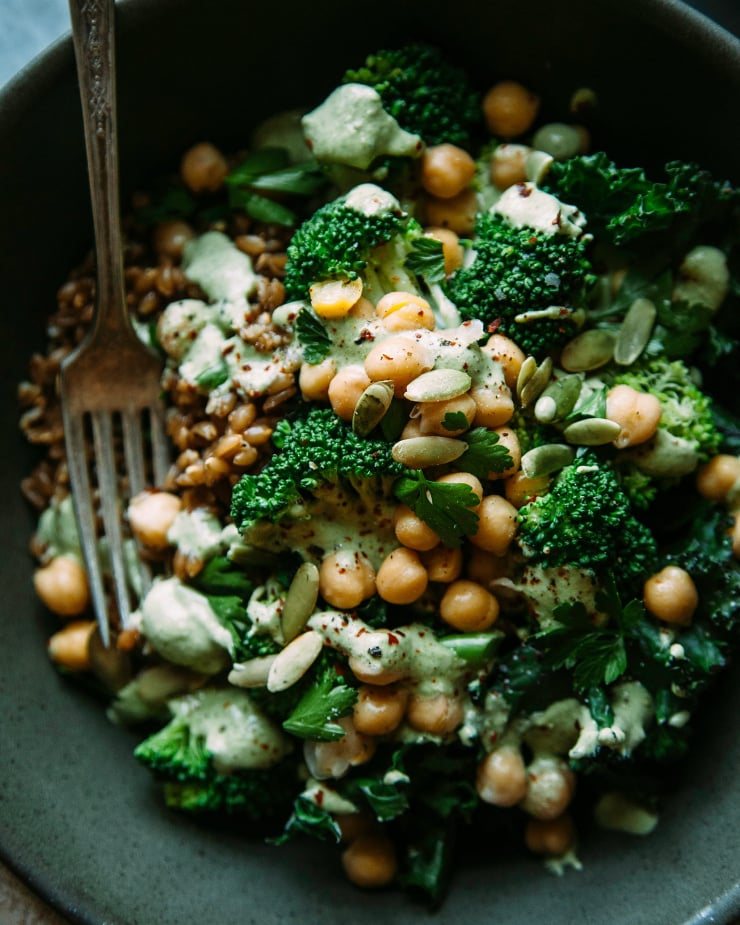 An up close shot of a green goddess bowl with broccoli, kale, chickpeas, farro, and a sage-green pepita lime sauce. The bowl is shown in a deep green matte bowl.