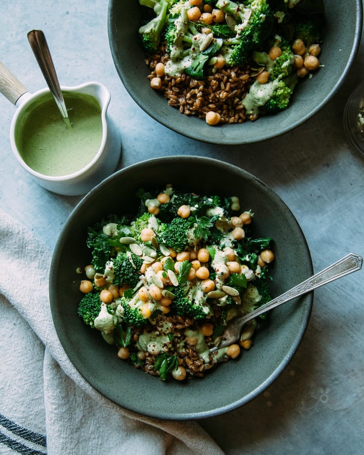An overhead shot of a green goddess bowl with broccoli, kale, chickpeas, farro, and a sage-green pepita lime sauce. The bowl is shown in a deep green matte bowl. A small pitcher of the sauce is shown nearby.