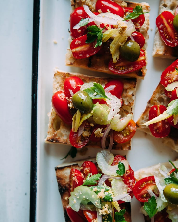 Overhead shot of vegan Bloody Mary bruschetta on a white enamelware tray with a black rim. The tomato-y bruschetta is garnished with olives, celery heart leaves and parsley.