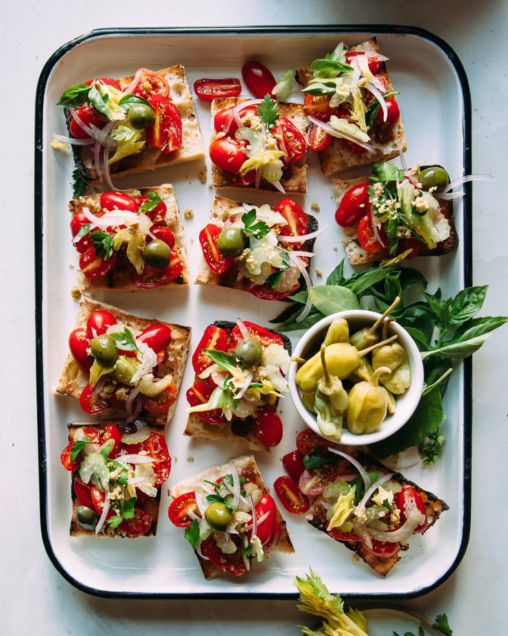 Overhead shot of vegan Bloody Mary bruschetta on a white enamelware tray with a black rim. The tomato-y bruschetta is garnished with olives, celery heart leaves and parsley.