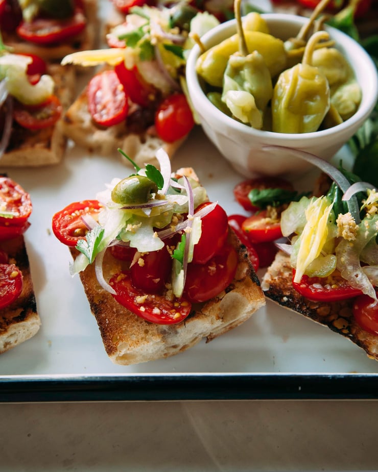 A slight 3/4 angle shot of cherry tomato bruschetta on crusty bread, all on a white tray. There is a bowl of pepperoncini peppers nearby. Part of a roundup of vegan appetizer recipes.