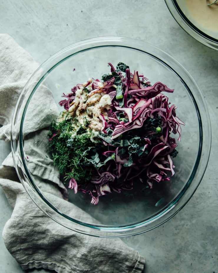 An overhead shot of shaved red cabbage, kale, chopped fresh dill, and a creamy dressing in a mixing bowl.