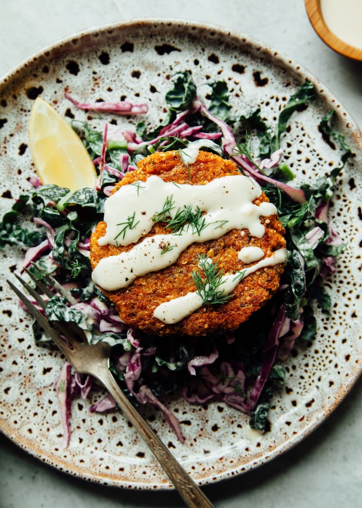 An overhead shot of a sweet potato cake pattie perched on top of a lemony slaw with a drizzle of tahini dressing. There is a lemon wedge to the side.