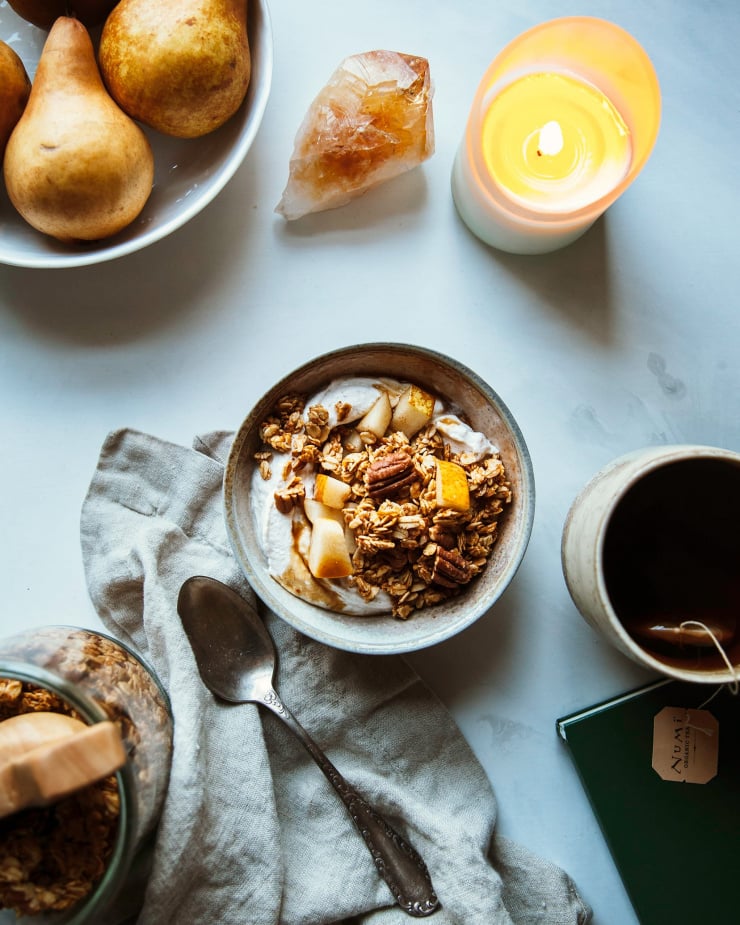 Overhead shot of sweet potato granola in a bowl with chopped pear and coconut milk granola. There is a lit candle to the side and a bowl of Bosc pears and a cup of tea.