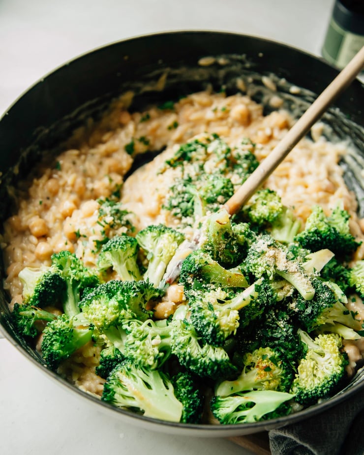 A 3/4 angle shot shows a wooden spoon stirring up a creamy lemon orzo with broccoli and chickpeas in a pot.