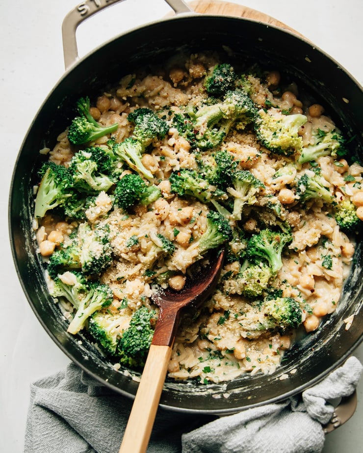 Overhead shot of creamy lemon orzo in a wide braiser pot with broccoli florets and chickpeas. There is a wooden spoon scooping up the orzo.