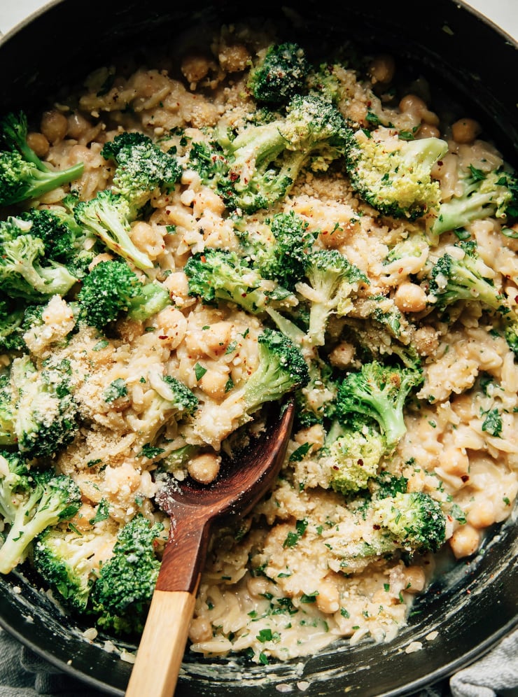 Up close overhead shot of creamy lemon orzo in a wide braiser pot with broccoli florets and chickpeas. There is a wooden spoon scooping up the orzo.