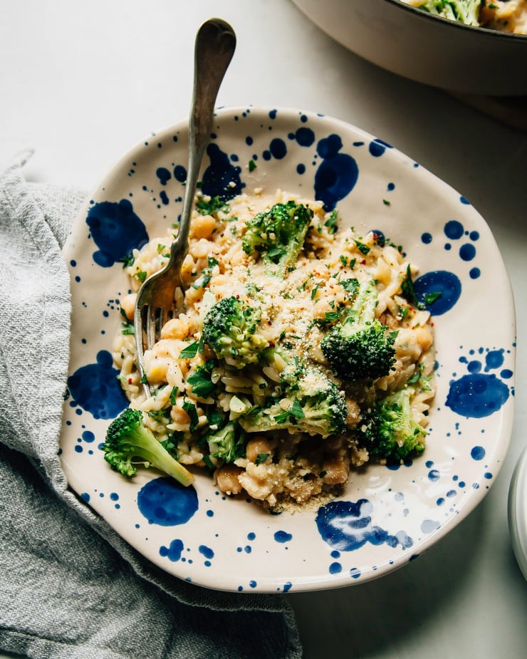 An overhead shot shows an individual bowl serving of creamy lemon orzo with chickpeas and broccoli. The bowl is ceramic and speckled with blue dots.