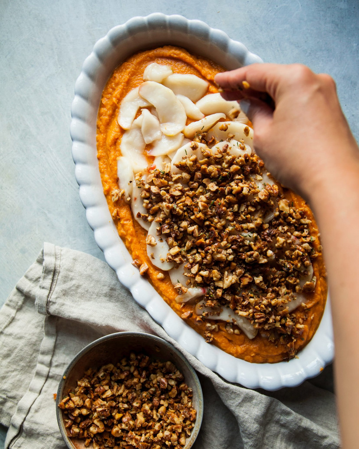 Image shows a hand sprinkling toasted walnuts on top of a sweet potato casserole.