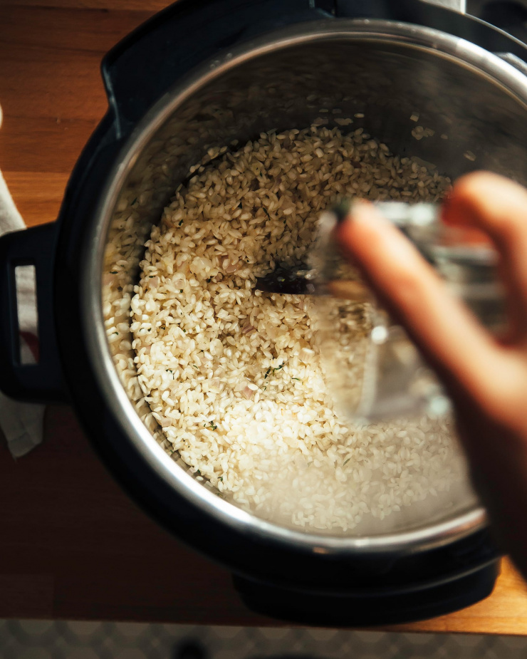 A hand is seen pouring white wine into a pot of toasting rice.