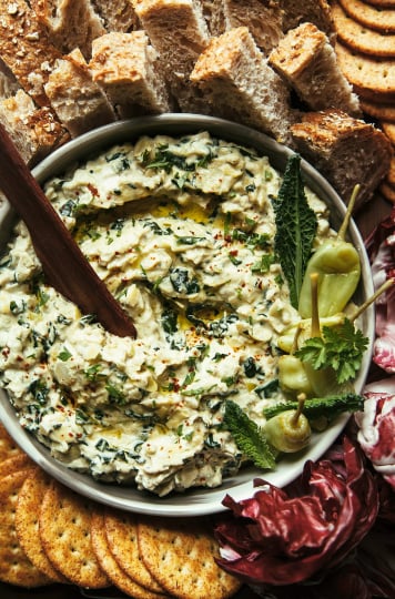 An overhead shot of a vegan appetizer recipe for a pepperoncini and artichoke dip with kale. The dip is surrounded by radicchio leaves, crackers, and pieces of bread.