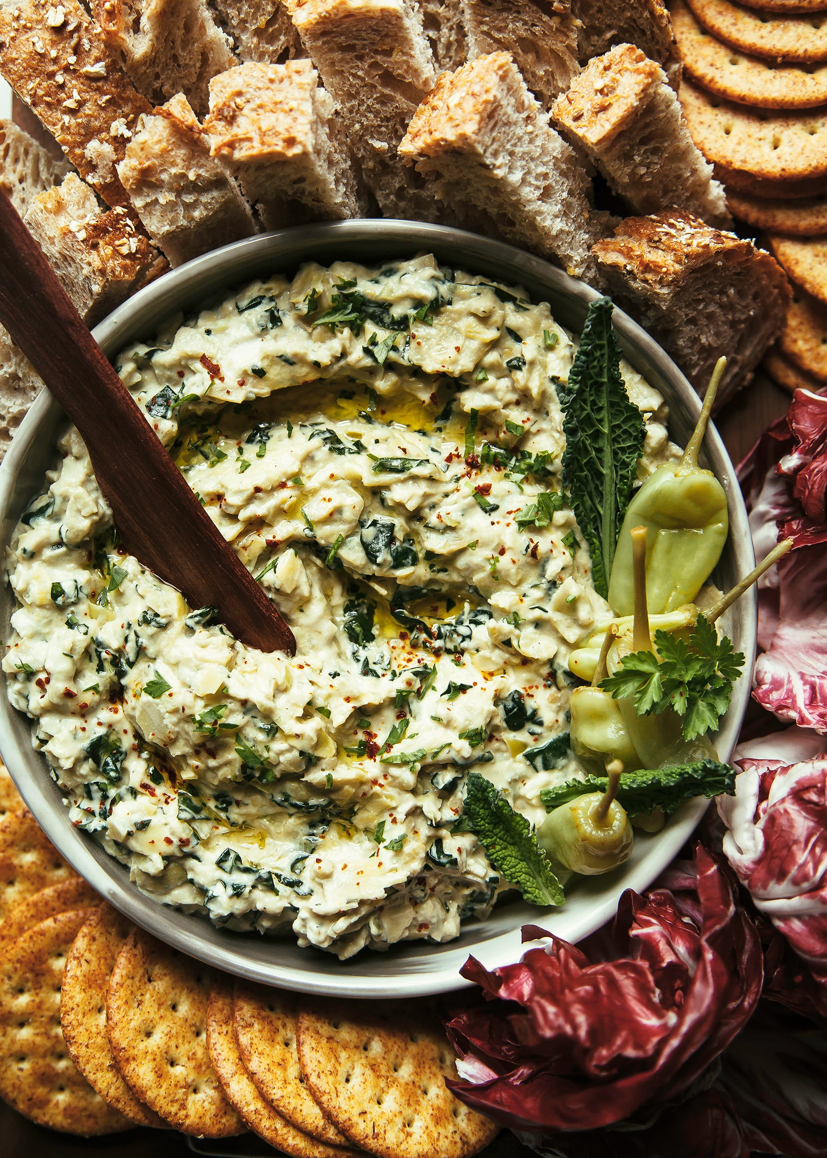 An overhead shot of a vegan appetizer recipe for a pepperoncini and artichoke dip with kale. The dip is surrounded by radicchio leaves, crackers, and pieces of bread.