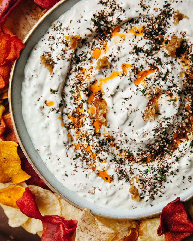 An up close, overhead shot of a creamy white dip with swirls of bright red chili oil and herbs on top. The bowl of dip is surrounded by multicoloured chips.