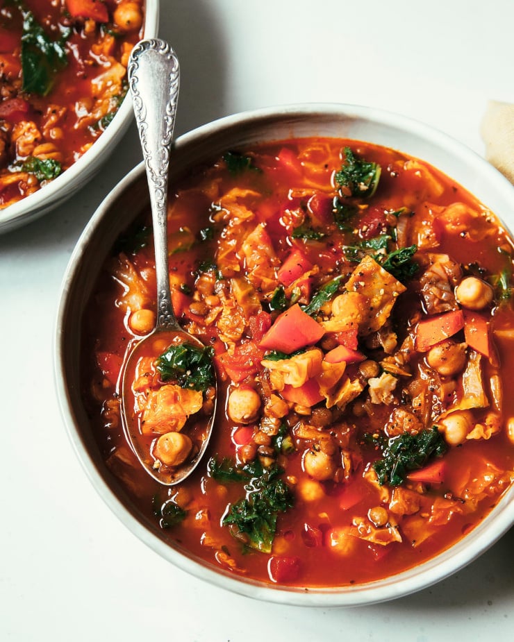 An overhead shot of a wide bowl of smoky chickpea, cabbage and kale soup. The soup is deep red and slightly chunky with little green bits of kale. The soup is photographed on a white background.