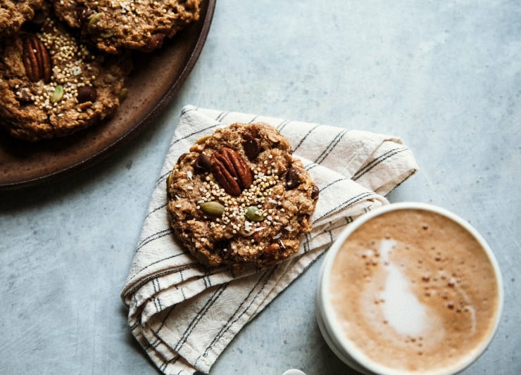 An overhead shot of a single cookie on a beige napkin alongside a frothy coffee.
