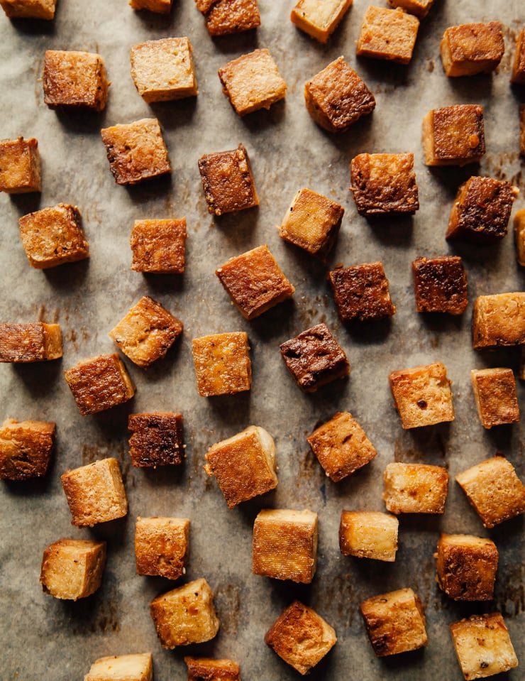 An overhead shot of crispy baked tofu cubes on a parchment lined baking sheet. The cubes of tofu are golden brown.