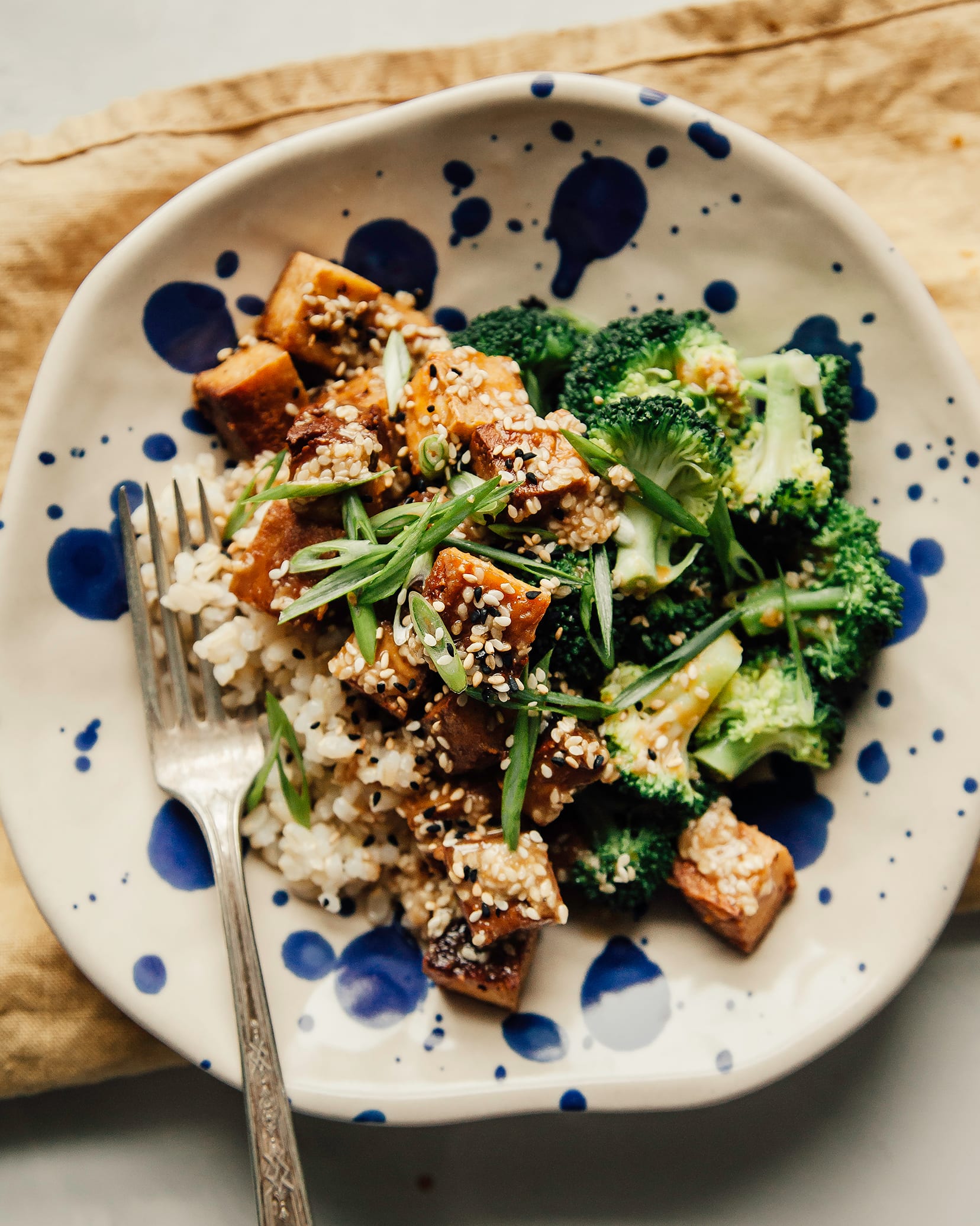 An overhead shot of a bowl containing crispy cubes of tofu, broccoli, cooked brown rice and sesame seeds.
