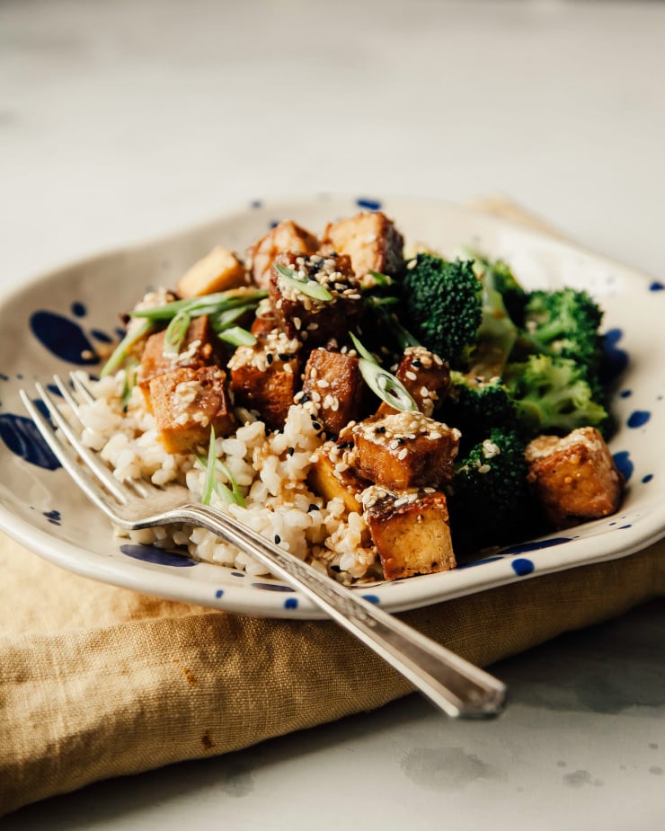 A head on show of a bowl filled with brown rice, crispy tofu cubes and cooked broccoli. A yellow linen napkin is seen to the side.