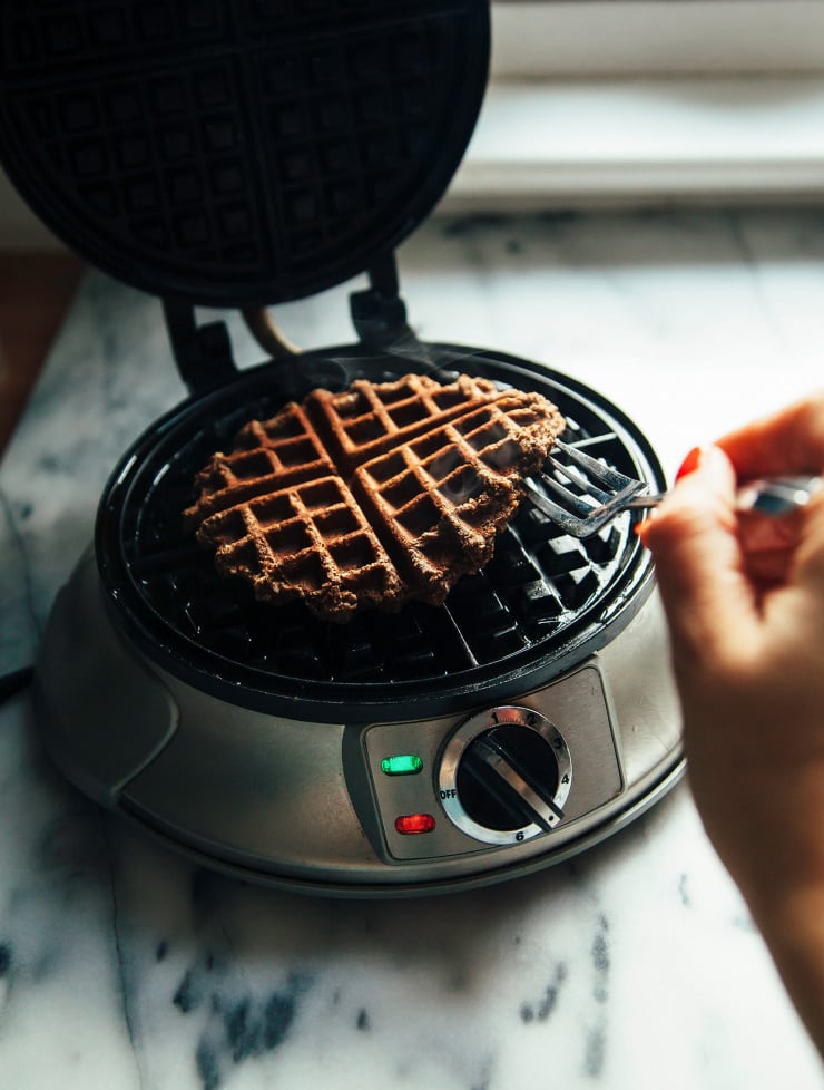 A hand lifting a hot waffle out of a waffle iron with a fork. There is steam billowing out.