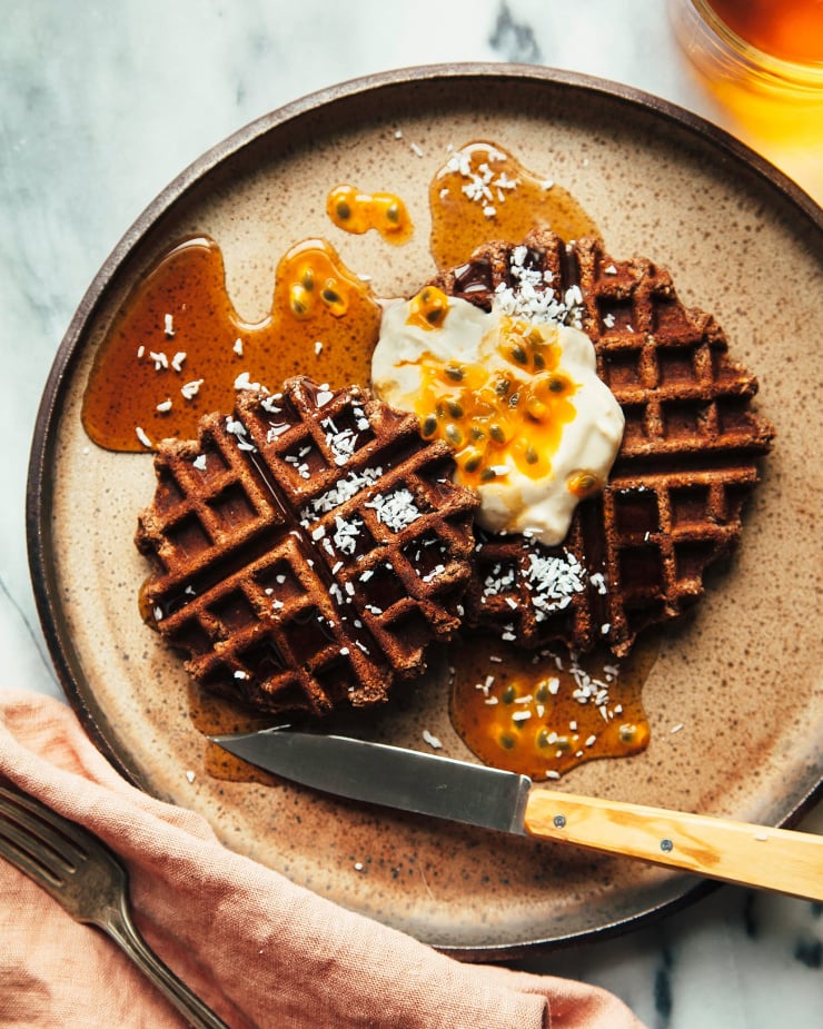 Overhead shot of buckwheat coconut waffles with coconut. Waffles are plated with a dollop of vegan yogurt and some passion fruit on top of a brown speckled plate.
