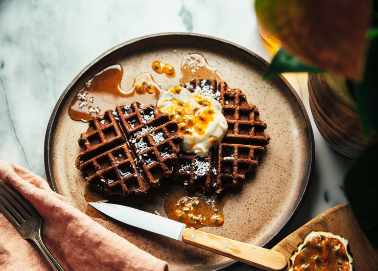Overhead shot of buckwheat coconut waffles with coconut. Waffles are plated with a dollop of vegan yogurt and some passion fruit on top of a brown speckled plate.