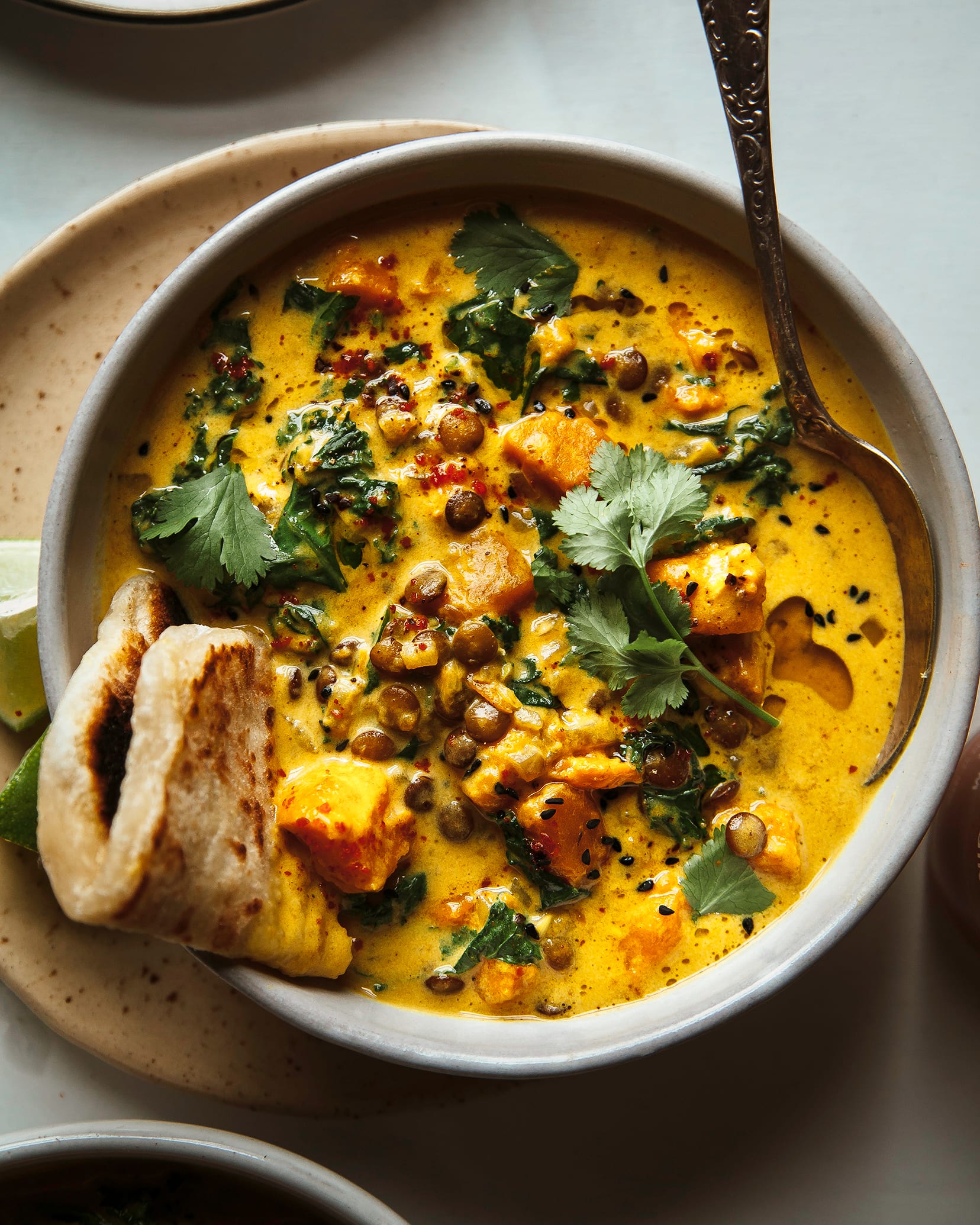 An overhead shot of a sweet potato and coconut milk stew in a bowl. The stew features brown lentils and kale and is topped with cilantro. A folded flatbread is tucked into one corner of the stew.