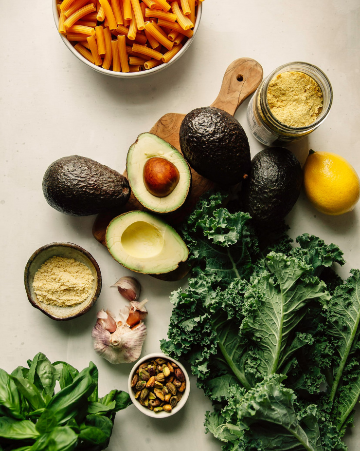 An overhead shot of ingredients for avocado pesto pasta with crispy kale on a white background.