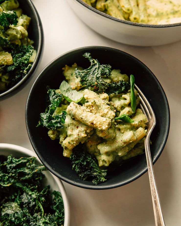An overhead shot of avocado pesto pasta in a dark green bowl with a yellow linen nearby. The pasta is garnished with crispy pieces of kale.